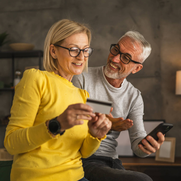 Mature or senior caucasian couple husband and wife shopping online sit at home husband hold mobile phone and woman hold and read information from credit card - Photo, Image