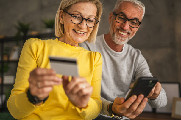 Mature or senior caucasian couple husband and wife shopping online sit at home husband hold mobile phone and woman hold and read information from credit card - Photo, Image