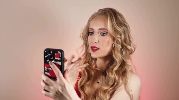 Une jeune femme en robe rouge prend un selfie avec de longs cheveux blonds et du maquillage. La fille sourit et regarde la caméra en gros plan - Séquence, vidéo