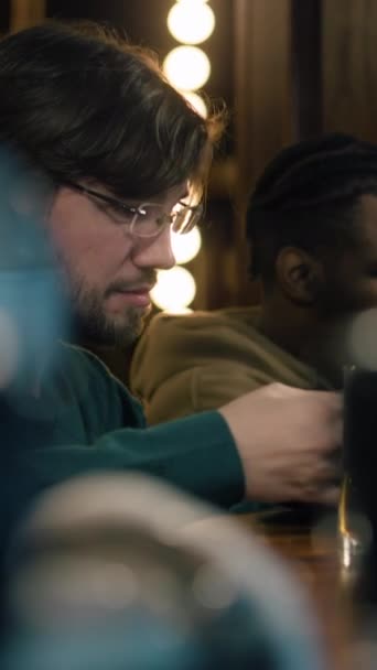 Vertikale Aufnahme eines Mannes mit Brille sitzt an der Theke, er trinkt, isst und unterhält sich mit einem afroamerikanischen Freund. Die Leute genießen ihren Wochenendabend in der Sportkneipe. Freunde verbringen Zeit zusammen in Bar - Filmmaterial, Video