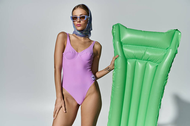 Modna kobieta w fioletowym stroju kąpielowym uderza w pozę obok zielonego dmuchanego materaca. - Zdjęcie, obraz