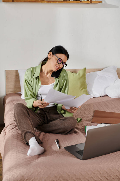 Женщина в повседневной одежде работает на своем ноутбуке в окружении бумаг на уютной кровати. - Фото, изображение