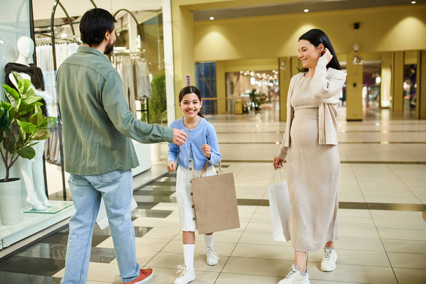 Ένας άνδρας και μια γυναίκα ψωνίζουν με την κόρη τους, απολαμβάνοντας μια εκδρομή το Σαββατοκύριακο σε ένα πολυσύχναστο εμπορικό κέντρο γεμάτο με καταστήματα και αγοραστές. - Φωτογραφία, εικόνα