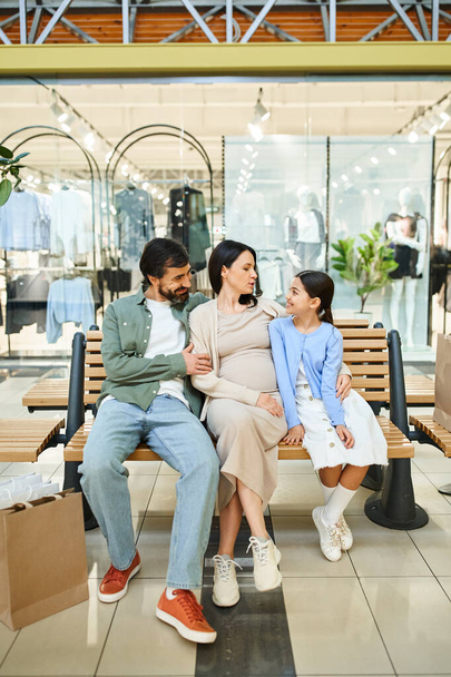 Μια ευτυχισμένη οικογένεια κάθεται σε ένα παγκάκι σε ένα πολυσύχναστο εμπορικό κέντρο, απολαμβάνοντας μια χαλαρωτική στιγμή μαζί κατά τη διάρκεια του Σαββατοκύριακου εκδρομή τους. - Φωτογραφία, εικόνα