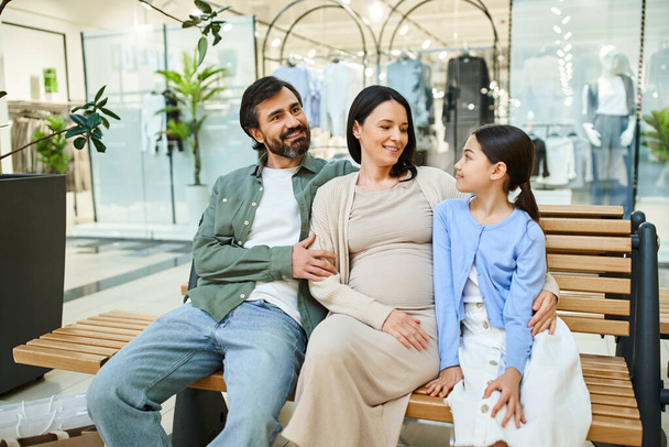 Eine schwangere Frau und ihre Tochter genießen einen Moment zusammen mit einem Mann auf einer Bank in einem Einkaufszentrum während eines Wochenendausflugs. - Foto, Bild