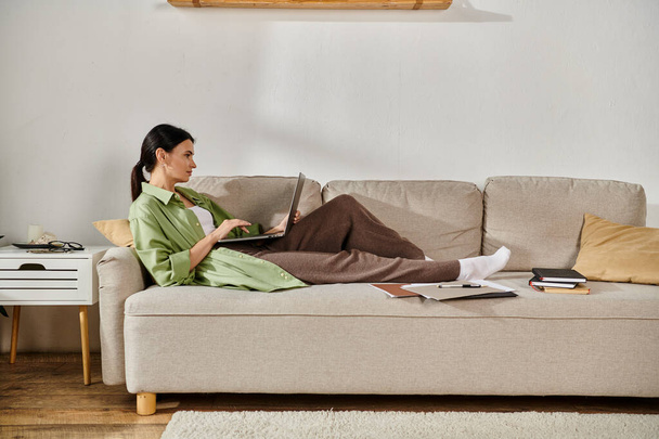 Eine geschäftige Frau in Freizeitkleidung arbeitet auf ihrem Laptop, während sie auf einer Couch sitzt. - Foto, Bild