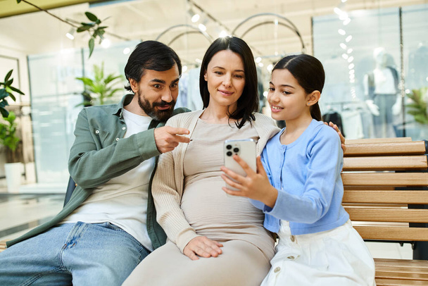 Μια οικογένεια κάθεται κοντά σε ένα παγκάκι σε ένα εμπορικό κέντρο, απορροφημένοι σε μια οθόνη του κινητού τηλεφώνου μαζί. - Φωτογραφία, εικόνα