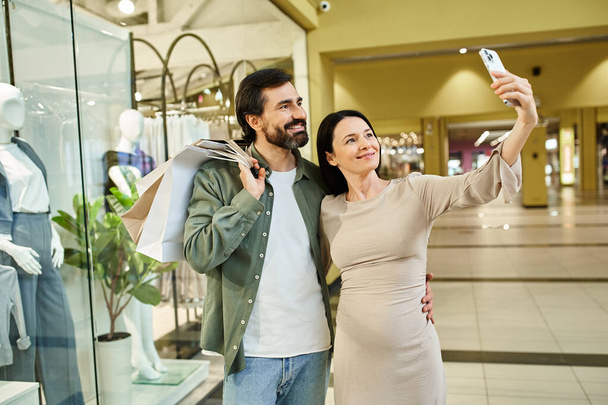 Um homem e uma mulher alegremente tiram uma selfie juntos enquanto fazem compras em um shopping. - Foto, Imagem