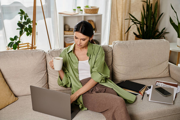Woman in casual attire, working on laptop with coffee in hand on cozy couch. - Photo, Image