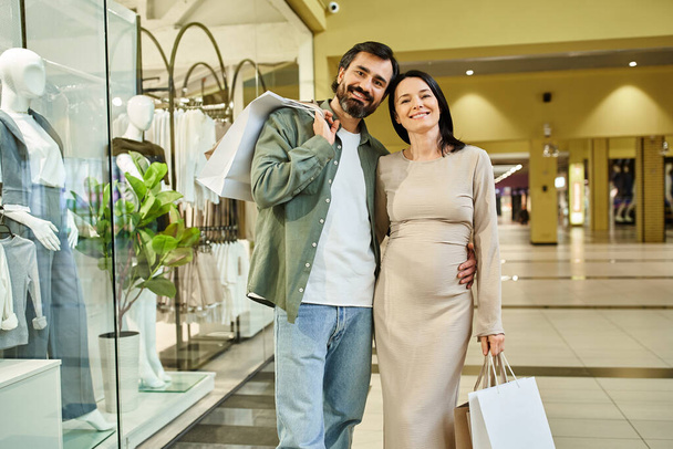 Embarazada pareja con entusiasmo sostiene bolsas de compras mientras explora un bullicioso centro comercial en una excursión de fin de semana. - Foto, imagen