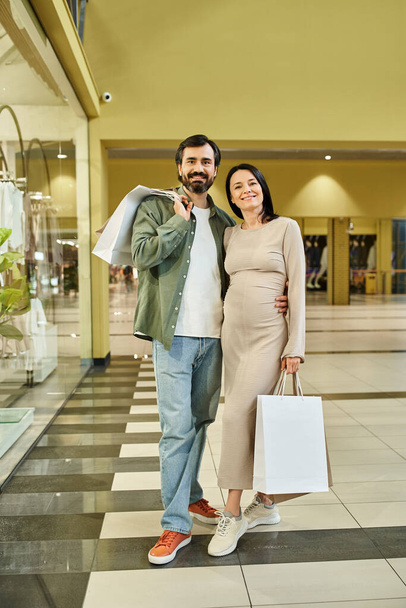 Ein schwangeres Paar hält fröhlich Einkaufstüten in einem belebten Einkaufszentrum und genießt einen gemeinsamen Wochenendausflug. - Foto, Bild