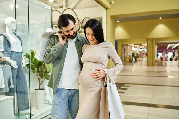 Μια έγκυος άντρας και γυναίκα ψωνίζουν ευτυχισμένοι μαζί σε ένα πολυσύχναστο εμπορικό κέντρο, απολαμβάνοντας το χρόνο τους ως μια αναπτυσσόμενη οικογένεια. - Φωτογραφία, εικόνα