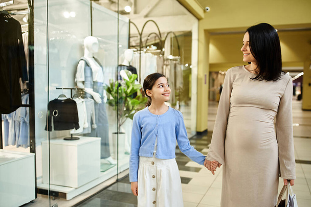 Μια έγκυος γυναίκα και η κόρη της εξερευνούν ένα εμπορικό κέντρο μαζί, απολαμβάνοντας μια οικογενειακή εμπειρία δεσίματος. - Φωτογραφία, εικόνα