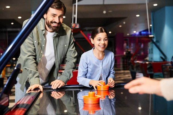 πατέρας και κόρη συμμετέχουν σε ένα φιλικό παιχνίδι χόκεϊ αέρα σε μια ζώνη τυχερών παιχνιδιών σε ένα εμπορικό κέντρο, δημιουργώντας μια ζωντανή και διασκεδαστική ατμόσφαιρα - Φωτογραφία, εικόνα