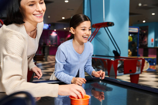 Eine fröhliche Mutter und ihre Tochter tauchen am Wochenende in ein temperamentvolles Air-Hockey-Match in einer Spielzone in einem Einkaufszentrum ein. - Foto, Bild