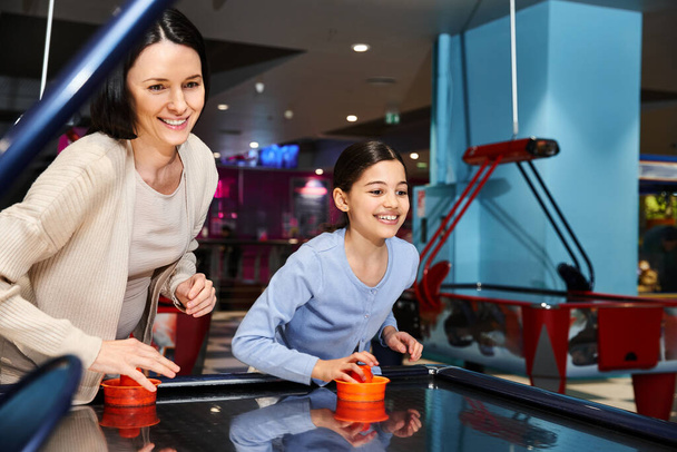 Μια ευτυχισμένη μητέρα και το παιδί παίζουν χόκεϊ αέρα σε μια ζώνη τυχερών παιχνιδιών εμπορικό κέντρο κατά τη διάρκεια μιας εξόρμησης Σαββατοκύριακο, απολαμβάνοντας την ποιότητα του χρόνου μαζί. - Φωτογραφία, εικόνα