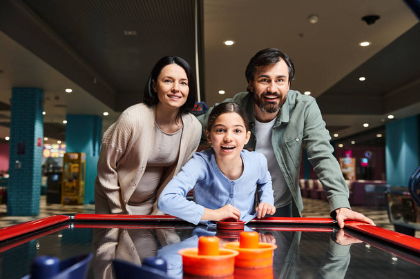 Μια ευτυχισμένη οικογένεια διαγωνίζεται σε ένα παιχνίδι χόκεϊ στον αέρα σε ένα arcade, γελώντας και απολαμβάνοντας ένα διασκεδαστικό Σαββατοκύριακο μαζί. - Φωτογραφία, εικόνα
