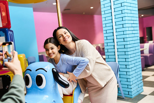 Una madre y su hija sonríen mientras disfrutan de un paseo en un carrusel de juguete en una zona de juego de centros comerciales el fin de semana. - Foto, imagen