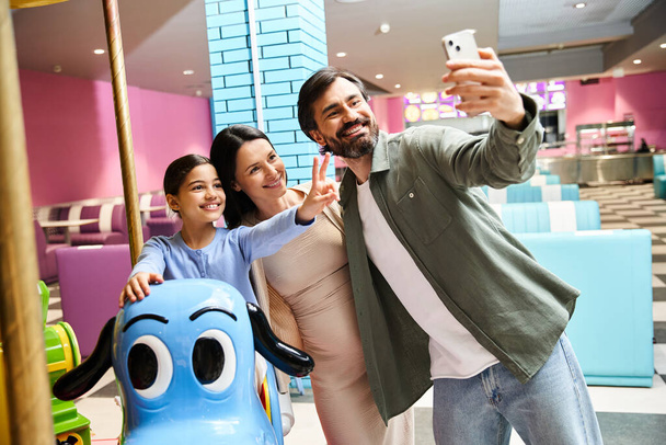 A joyful family smiles while taking a selfie in front of a carousel toy at a malls gaming zone during the weekend. - Photo, Image