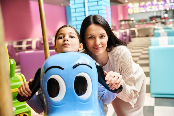 Eine fröhliche Frau und ein Kind posieren fröhlich zusammen neben einem bunten Karussell-Spielzeug in einem Einkaufszentrum. - Foto, Bild
