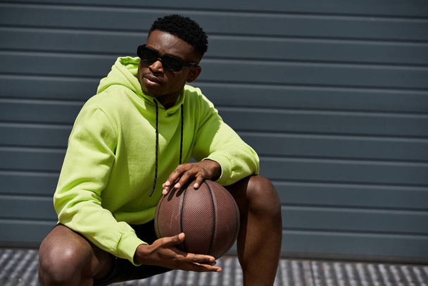 Fiatal afro-amerikai férfi zöld kapucnis pulcsiban, kosárlabdával a kezében. - Fotó, kép