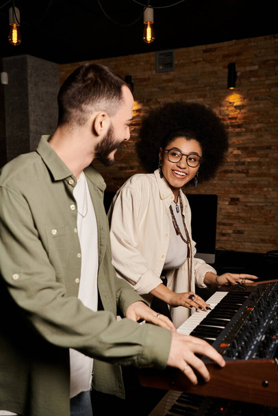 Ένας άντρας και μια γυναίκα στέκονται δίπλα σε ένα πληκτρολόγιο σε ένα στούντιο ηχογράφησης, βυθισμένοι στη δημιουργία μουσικής για την πρόβα της μπάντας τους.. - Φωτογραφία, εικόνα