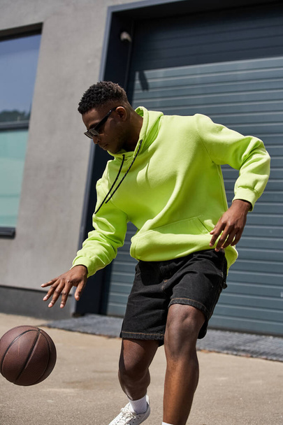 Schöner afroamerikanischer Mann trägt neonfarbenen Kapuzenpulli und kurze Hosen und hält Basketball in der Hand. - Foto, Bild