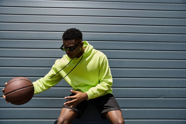 Afrikai származású fiatalember élénk zöld kapucnis pulóverben, kosárlabdával a kezében.. - Fotó, kép