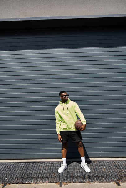 ガレージの前にバスケットボールを保持するファッショナブルな服装のハンサムな若いアフリカ系アメリカ人男性. - 写真・画像