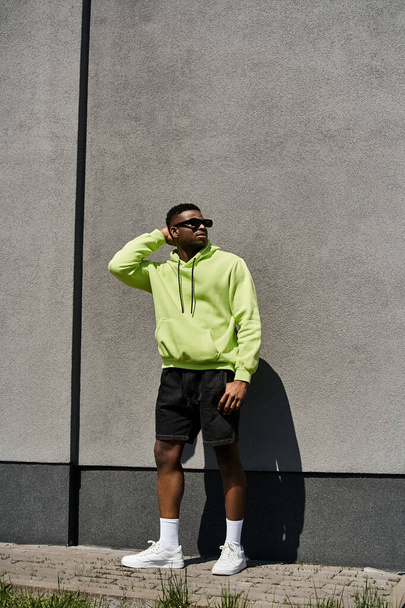 Stijlvolle man in neon hoodie en korte broek leunend tegen een muur. - Foto, afbeelding