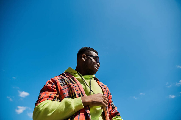 Elegante hombre afroamericano en chaqueta brillante se encuentra bajo el cielo azul claro. - Foto, imagen