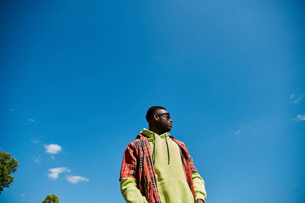 Ένας μοντέρνος Αφροαμερικανός που στέκεται σε ένα χωράφι κάτω από έναν γαλάζιο ουρανό.. - Φωτογραφία, εικόνα