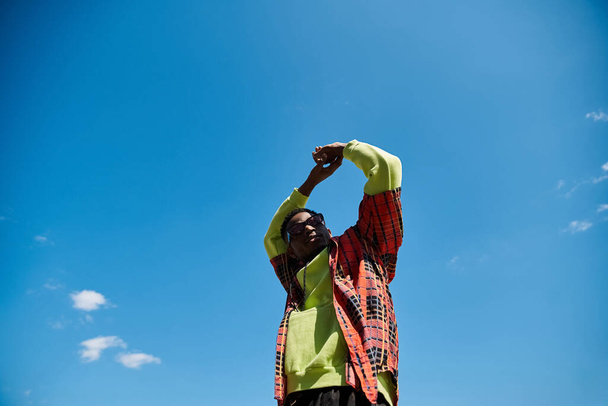 Ένας μοντέρνος νεαρός Αφροαμερικανός που στέκεται σε ένα χωράφι κάτω από έναν γαλάζιο ουρανό.. - Φωτογραφία, εικόνα
