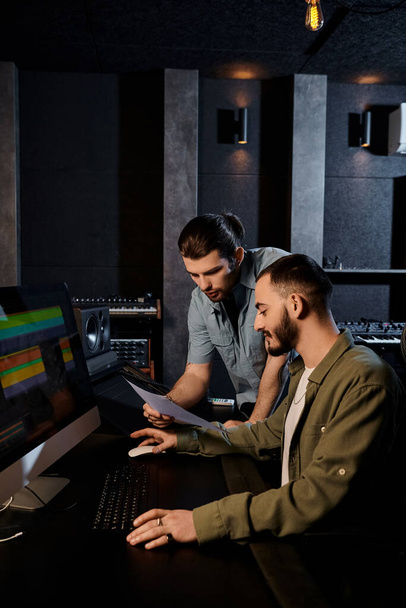 Δύο άνδρες σε ένα στούντιο ηχογράφησης επικεντρώνονται έντονα στην οθόνη του υπολογιστή τους κατά τη διάρκεια μιας πρόβας μουσικής μπάντας. - Φωτογραφία, εικόνα