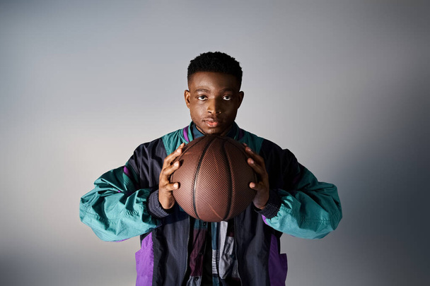 Jóképű afroamerikai férfi divatos öltözékben, szürke háttér előtt tartva a kosárlabdát. - Fotó, kép