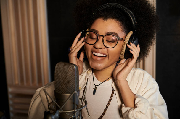 Uma mulher com óculos e cabelo afro canta apaixonadamente em um microfone durante um ensaio da banda de música em um estúdio de gravação. - Foto, Imagem