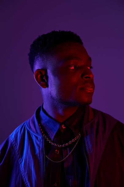 Handsome African American man in fashionable jacket and chain against vivid purple backdrop. - Photo, Image
