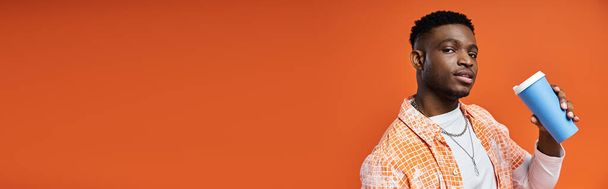 Fešák Afroameričan ve stylovém oblečení drží kávu na oranžovém pozadí. - Fotografie, Obrázek