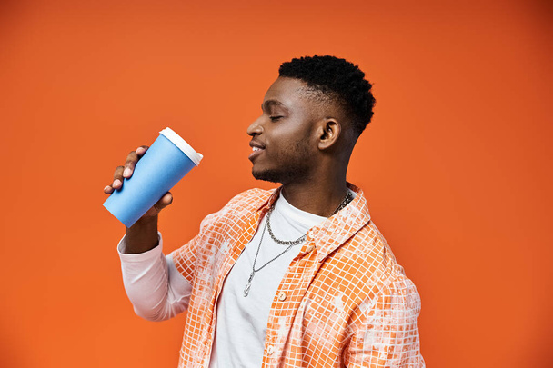 Knappe Afro-Amerikaanse man drinkt uit blauwe beker tegen levendige oranje achtergrond. - Foto, afbeelding