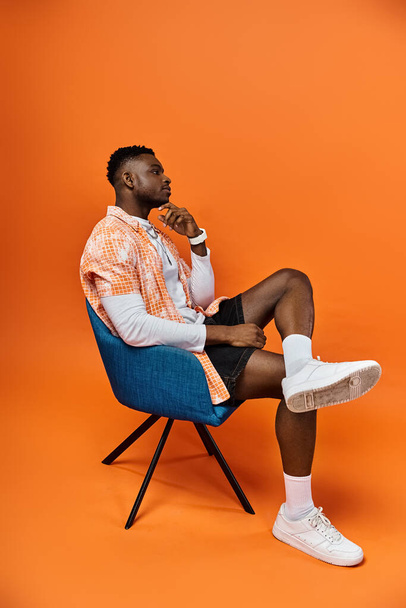 Модный афроамериканец сидит в стильном кресле на ярко-оранжевом фоне. - Фото, изображение