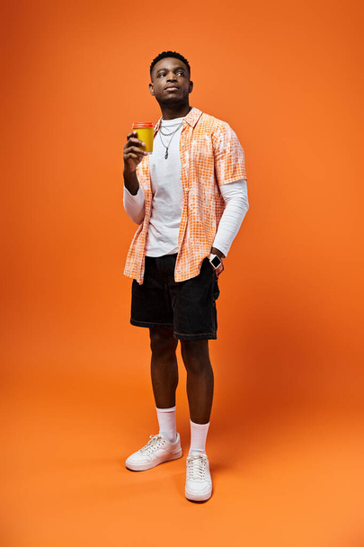 Bell'uomo afroamericano in abiti alla moda che tiene un drink davanti a uno sfondo arancione brillante. - Foto, immagini
