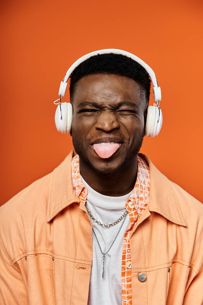 Homme afro-américain élégant portant des écouteurs avec la langue qui sort dans un geste amusant et ludique. - Photo, image