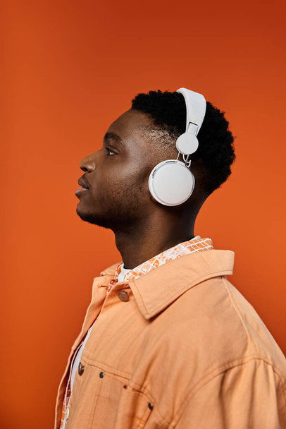 明るいオレンジ色の背景の前にヘッドフォンで音楽を楽しんでいるファッショナブルな若いアフリカ系アメリカ人男性. - 写真・画像