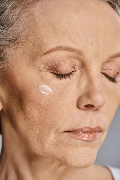 Μια ώριμη γυναίκα απλώνει κρέμα στο πρόσωπό της για μια αναζωογονητική ρουτίνα φροντίδας του δέρματος. - Φωτογραφία, εικόνα