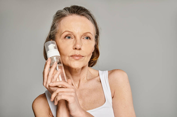 Una mujer madura en traje acogedor sosteniendo una botella de limpiador facial. - Foto, Imagen
