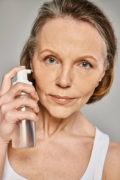 Una mujer madura y atractiva con un atuendo cómodo sostiene una botella de producto para el cuidado de la piel. - Foto, Imagen