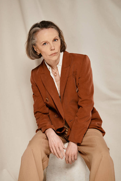 Stilvolle reife Frau in braunem Blazer und brauner Hose sitzt bequem auf einem Hocker. - Foto, Bild