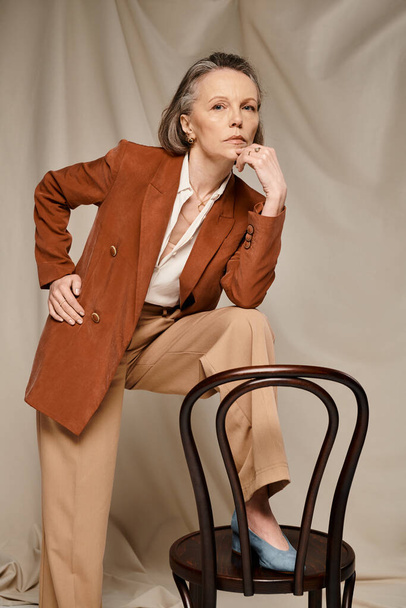 Mature woman poses actively in tan blazer and pants on chair. - Photo, Image
