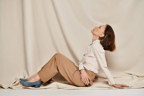 Женщина в белой рубашке и коричневых штанах изящно сидит на кровати. - Фото, изображение