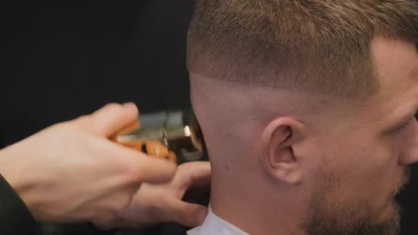 Kampaaja ajelee nuoren miehen napa sähköparranajokoneella parturissa lähikuva. Barber tekee tyylikkään kampauksen asiakkaalle kampaamossa - Materiaali, video
